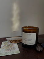 An amber glass jar candle with a white label reading 'dreamweaver' and 'heartsong' sits on a dark wood surface, surrounded by a tarot card, crystals, and a flower of life wood carving.