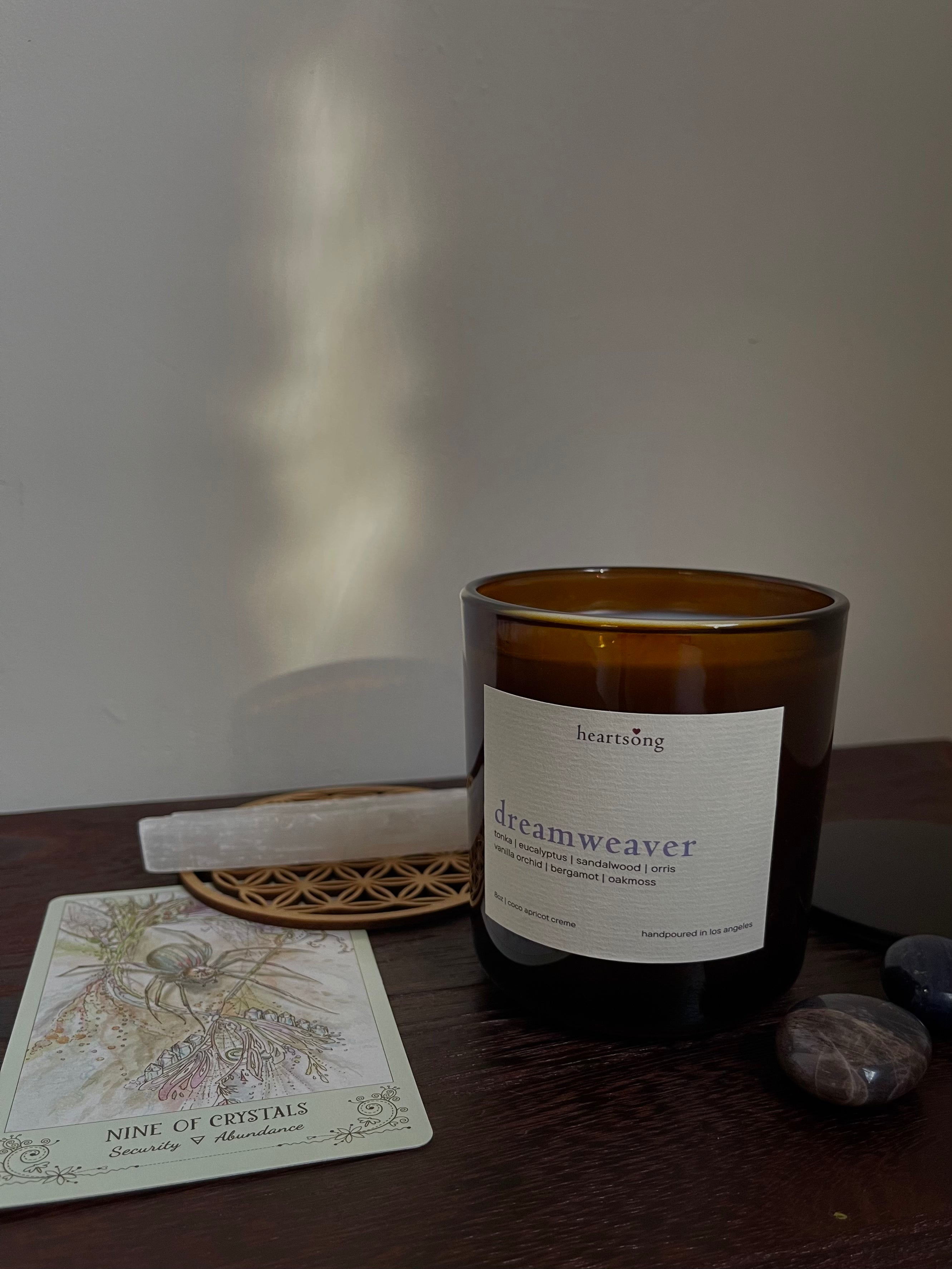An amber glass jar candle with a white label reading 'dreamweaver' and 'heartsong' sits on a dark wood surface, surrounded by a tarot card, crystals, and a flower of life wood carving.