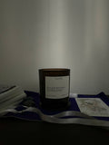 An amber glass jar candle with a white label reading 'dreamweaver' sits on a dark surface draped with a violet cloth and a white ribbon, surrounded by cards and a small stone against a plain wall.