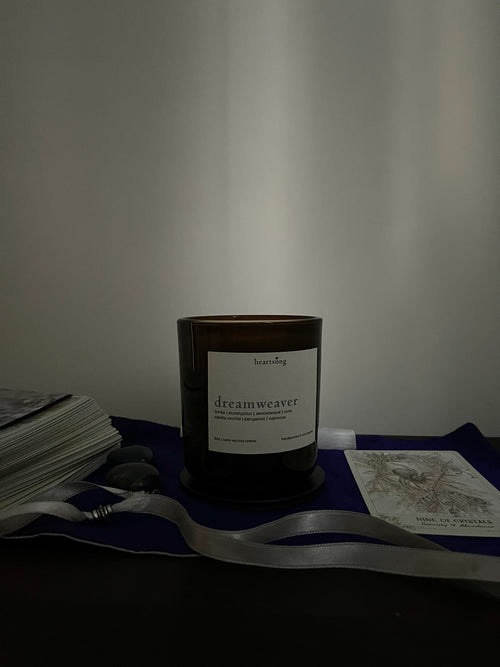 An amber glass jar candle with a white label reading 'dreamweaver' sits on a dark surface draped with a violet cloth and a white ribbon, surrounded by cards and a small stone against a plain wall.