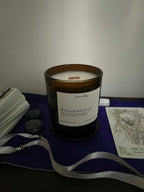An amber glass jar candle with a white label reading 'heartsong dreamweaver' sits on a dark surface draped with a purple cloth, surrounded by a white card deck, tarot card, gray crystals, and a white ribbon.