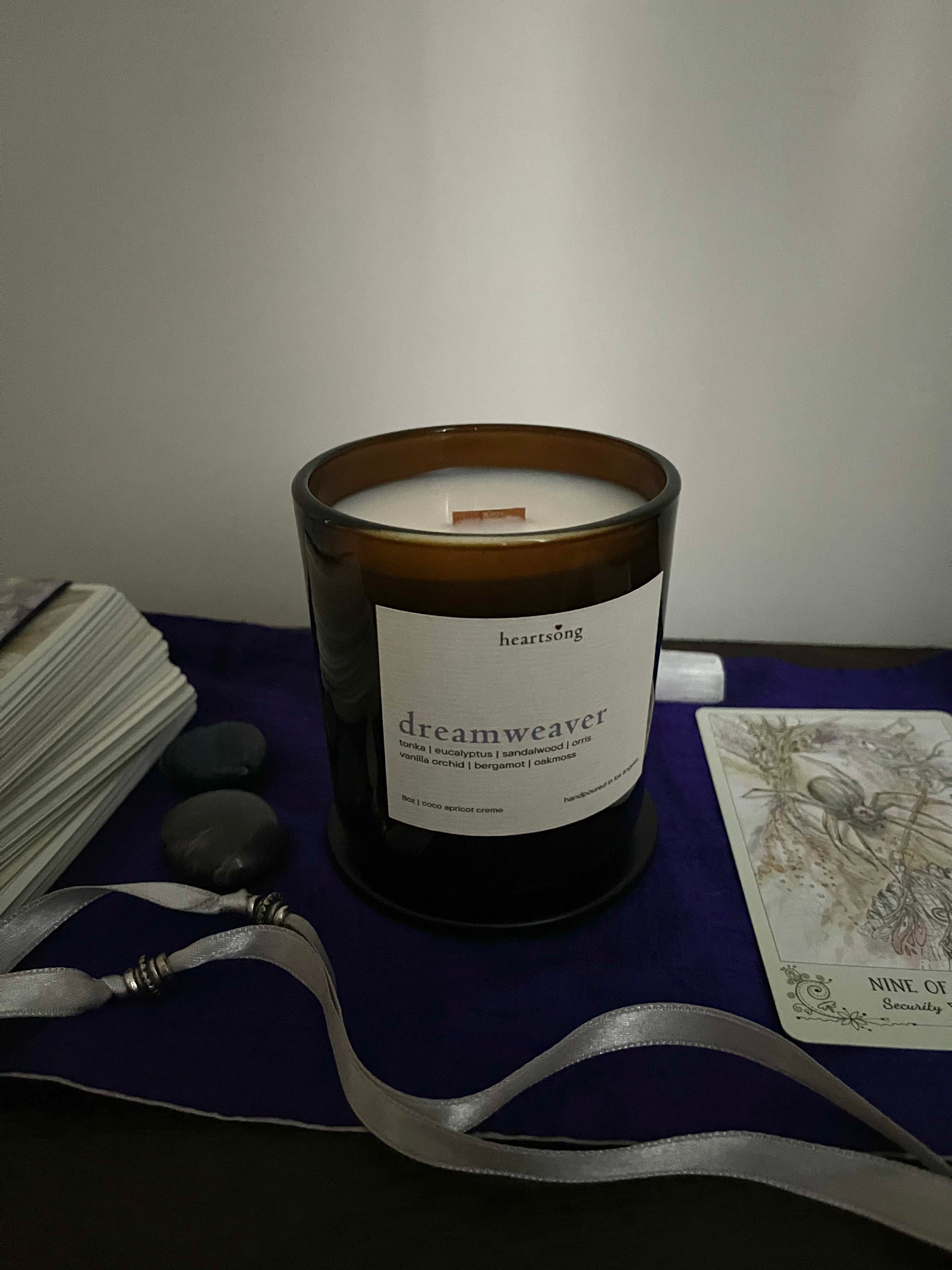 An amber glass jar candle with a white label reading 'heartsong dreamweaver' sits on a dark surface draped with a purple cloth, surrounded by a white card deck, tarot card, gray crystals, and a white ribbon.