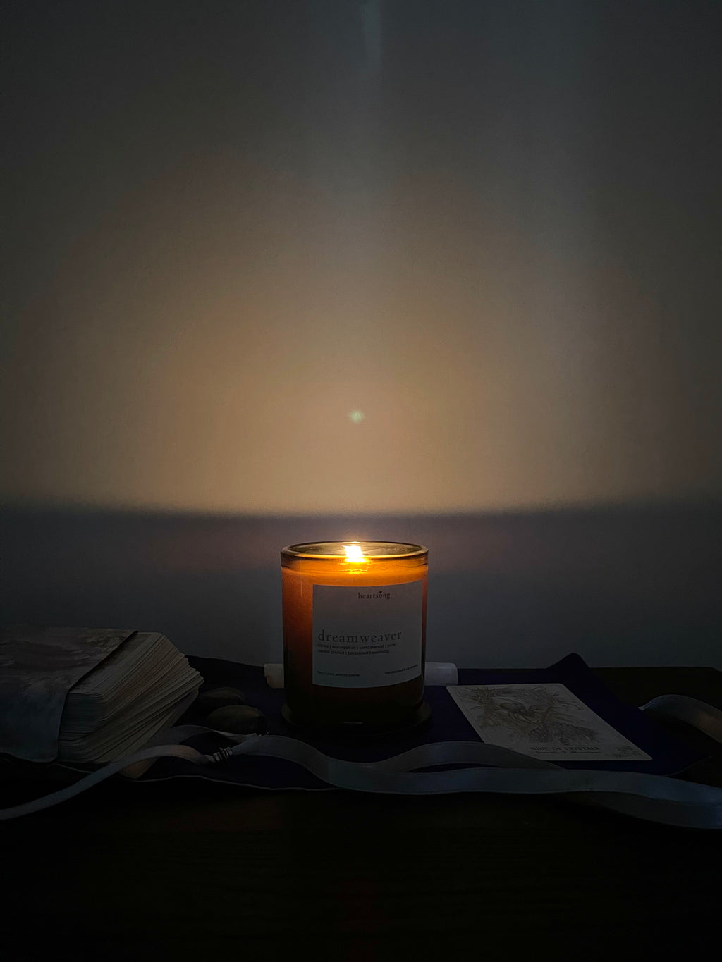 A lit candle in a dark amber glass jar sits on a dark surface against a dim wall, casting a warm glow. A white label is visible on the front, and nearby are stacked tarot cards, crystals and a ribbon.