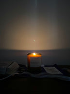 A lit candle in a dark amber glass jar sits on a dark surface against a dim wall, casting a warm glow. A white label is visible on the front, and nearby are stacked tarot cards, crystals and a ribbon.