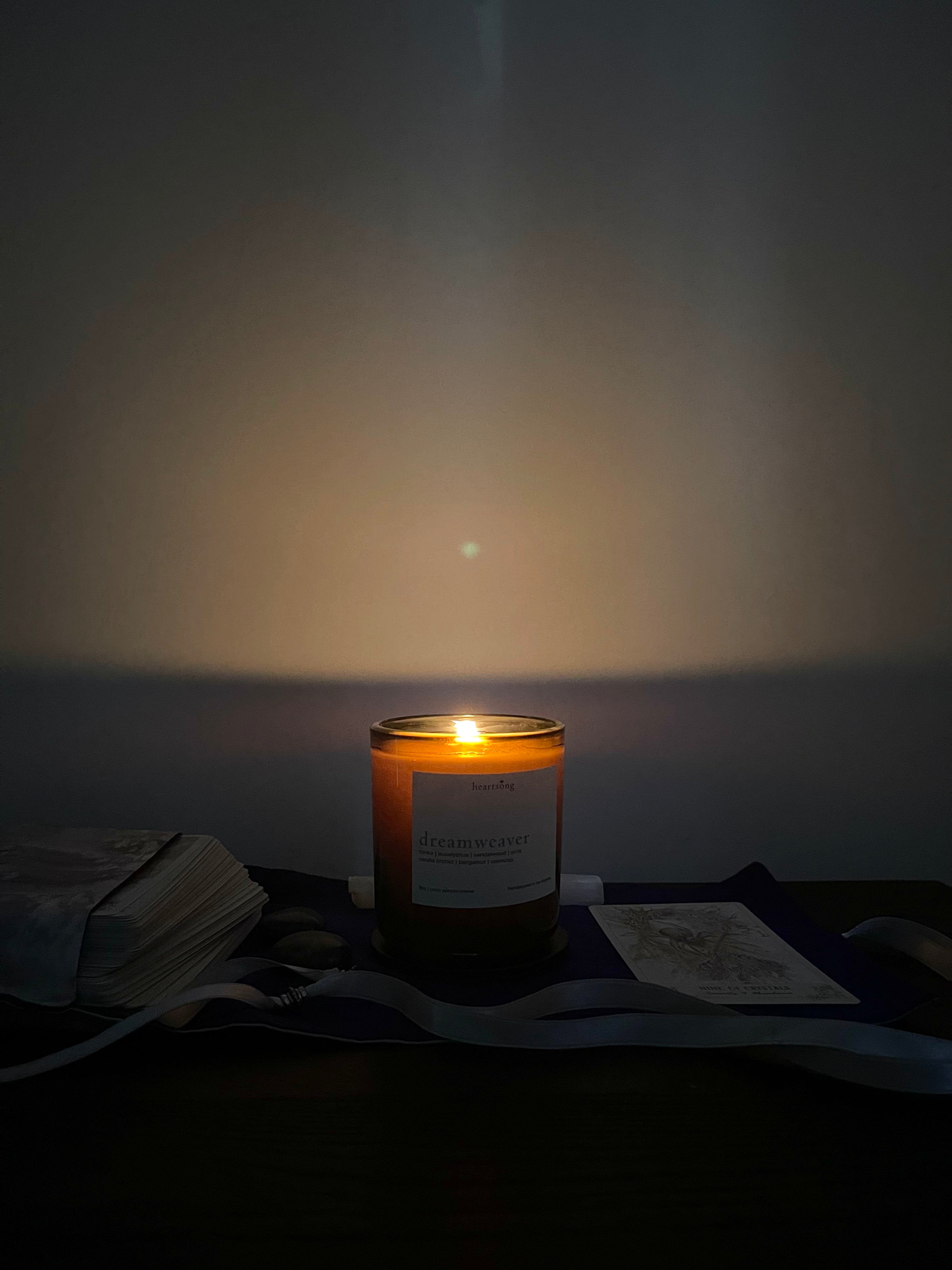 A lit candle in a dark amber glass jar sits on a dark surface against a dim wall, casting a warm glow. A white label is visible on the front, and nearby are stacked tarot cards, crystals and a ribbon.