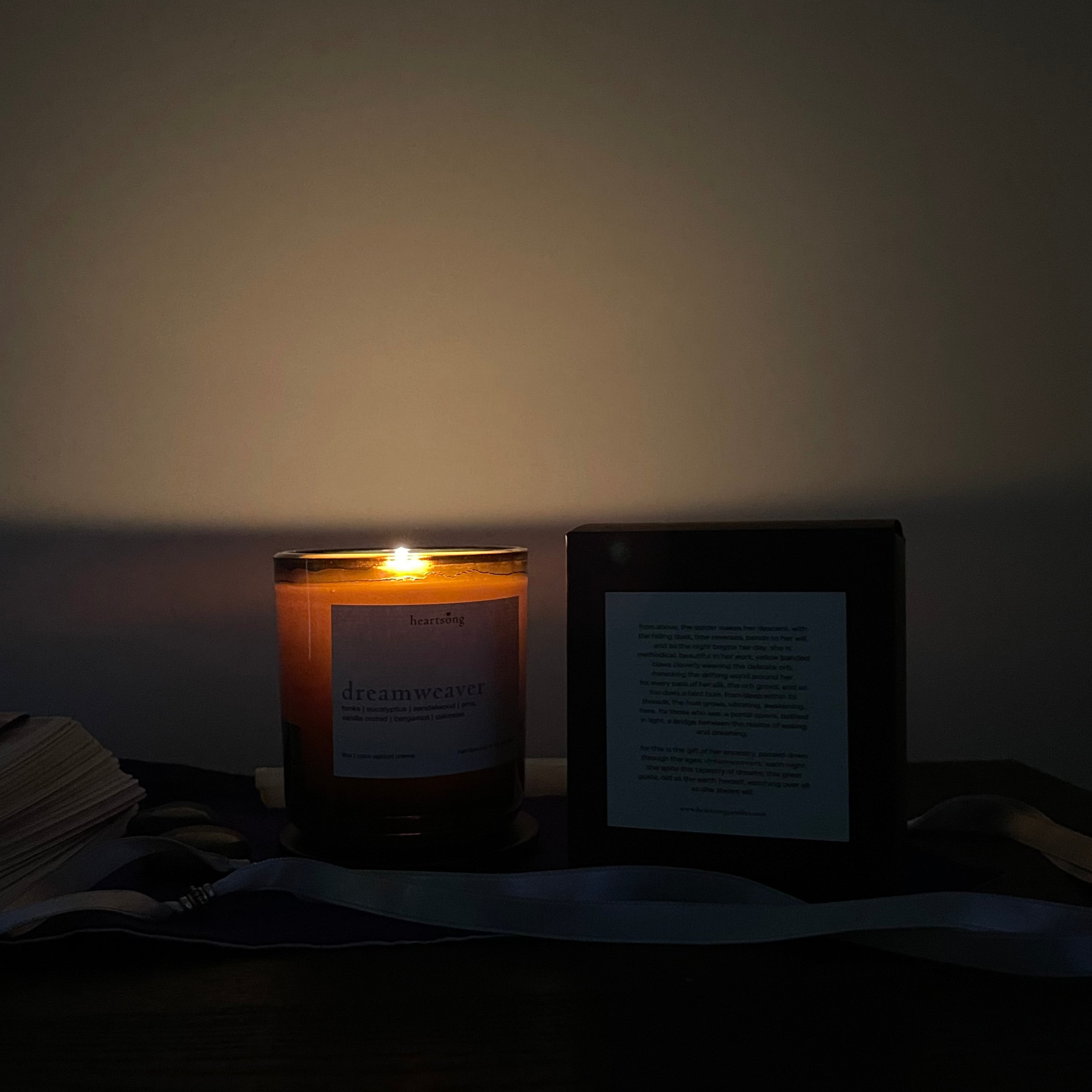 A lit amber glass candle and a deep brown candle box displaying a typed story labeled on the back, placed on a dark table against a dimly lit wall.