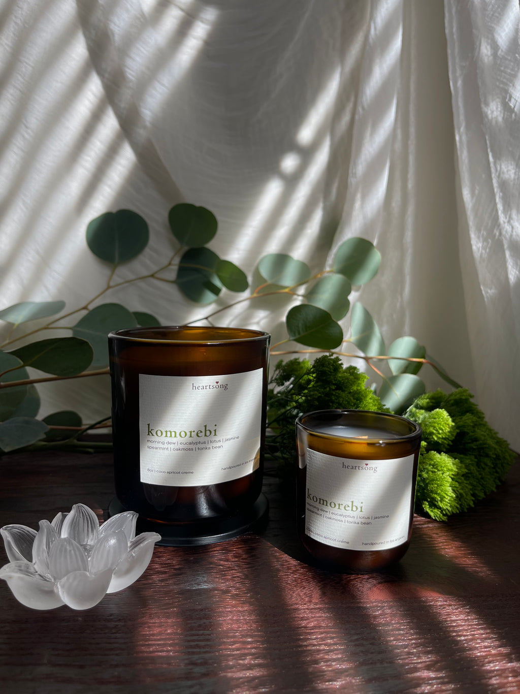 Two amber glass candles with white labels reading ‘komorebi', one small and one larger, styled on a dark wooden surface with eucalyptus leaves, moss, and a translucent glass lotus figurine under draped white fabric with soft window shadows.