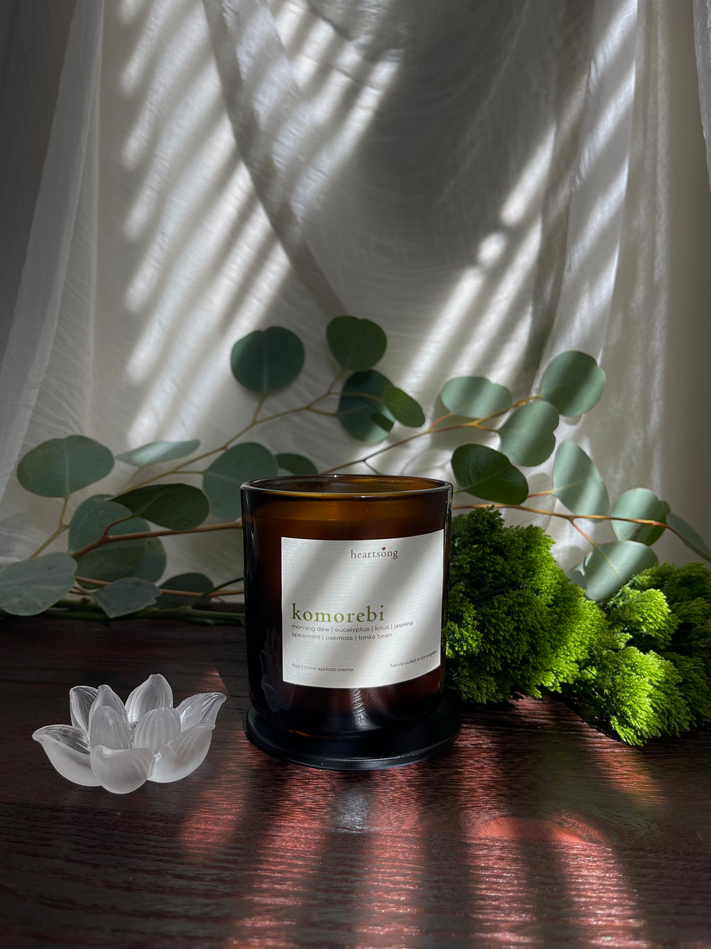 An amber glass jar candle with a white label reading 'komorebi' and ‘heartsong' at the top, styled on a dark wooden surface with eucalyptus leaves, green moss, a translucent glass lotus figurine, and draped white fabric with window shadow patterns across the scene.