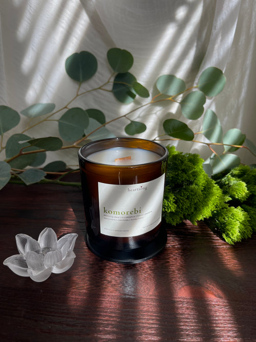 An amber glass jar candle with a white label reading ‘komorebi' sits on a dark wooden surface, styled with eucalyptus leaves, green moss, a clear lotus-shaped figurine, and draped white fabric with leaf shadows.