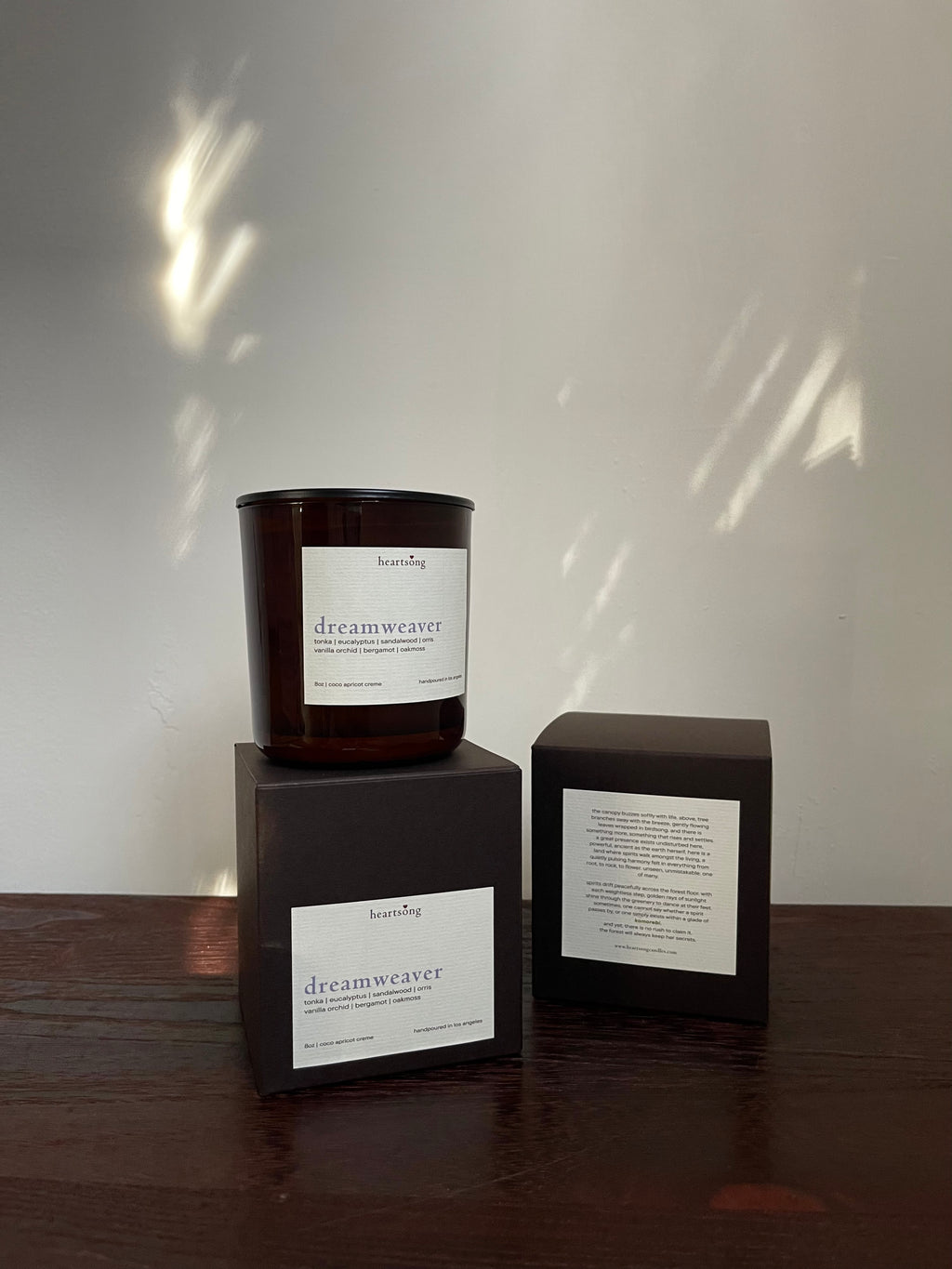A dark amber glass candle jar with a white label reading 'dreamweaver' sits on top of a dark brown box, alongside a second matching box showing the back panel, set on a wooden surface against a light wall with dappled sunlight.