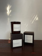A dark amber glass candle jar with a white label reading 'dreamweaver' sits on top of a dark brown box, alongside a second matching box showing the back panel, set on a wooden surface against a light wall with dappled sunlight.