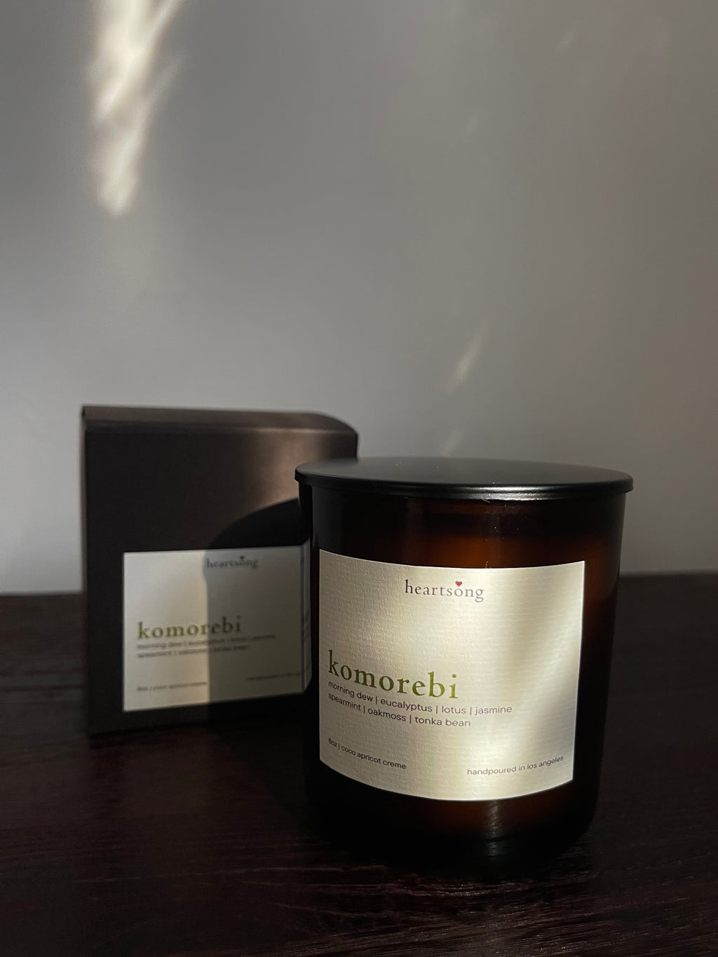 An amber glass jar candle labeled ‘komorebi' with a dark lid sits on a dark wooden surface, with its matching dark box behind it against a softly lit neutral wall.