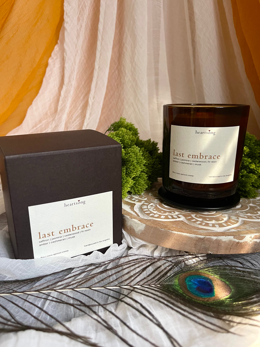 An amber glass candle jar labeled 'last embrace' by heartsong, shown beside its dark brown box with a matching label. The setup includes draped cream fabric, mustard yellow fabric, green moss, a carved wooden lotus stand, and a peacock feather in the foreground.