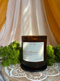 An amber glass jar candle with a white label reading ‘heartsong' and 'last embrace' sits on a patterned wooden lotus tray surrounded by green foliage, with draped cream fabric and mustard-yellow drapery in the background.