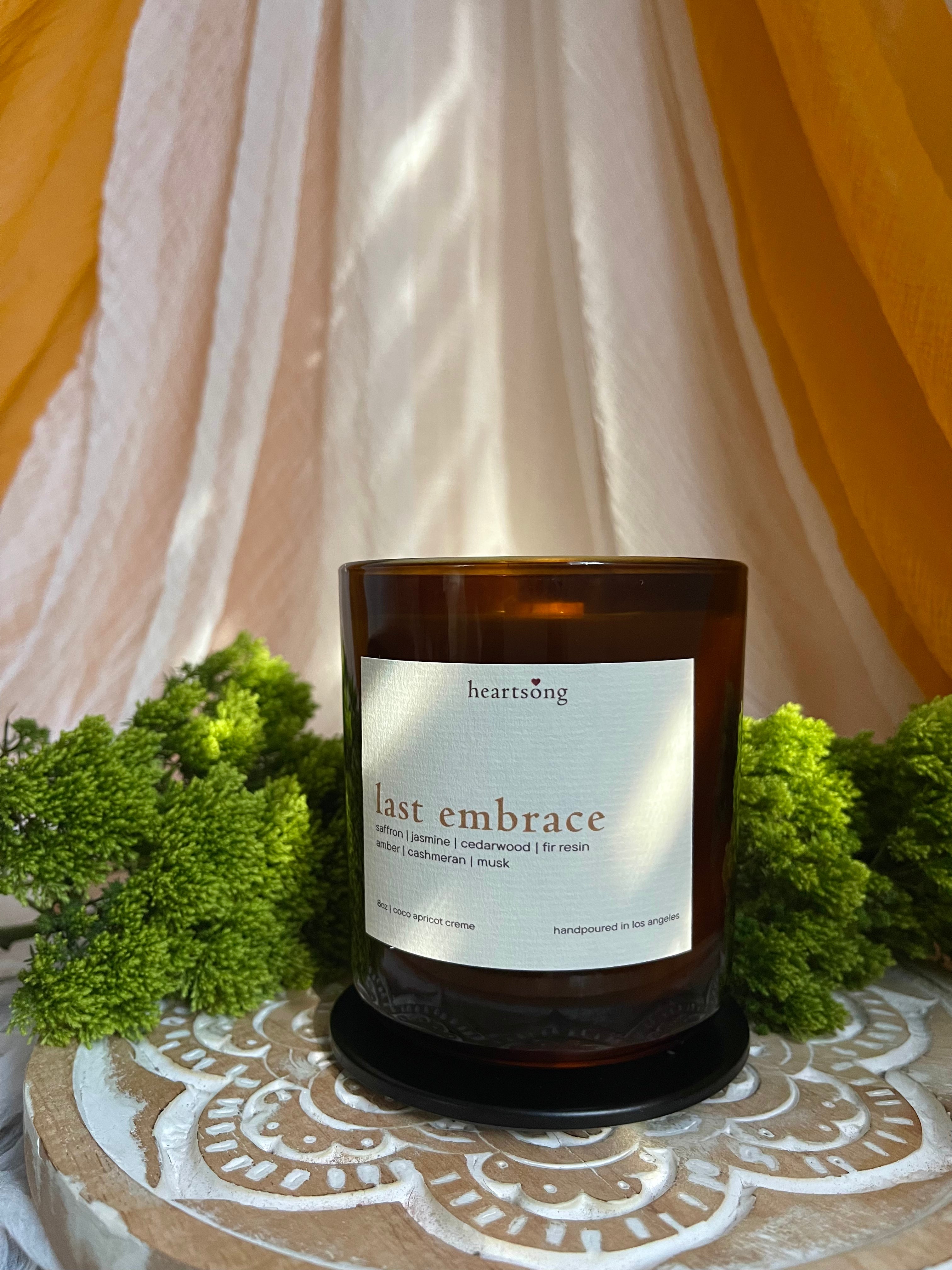 An amber glass jar candle with a white label reading ‘heartsong' and 'last embrace' sits on a patterned wooden lotus tray surrounded by green foliage, with draped cream fabric and mustard-yellow drapery in the background.