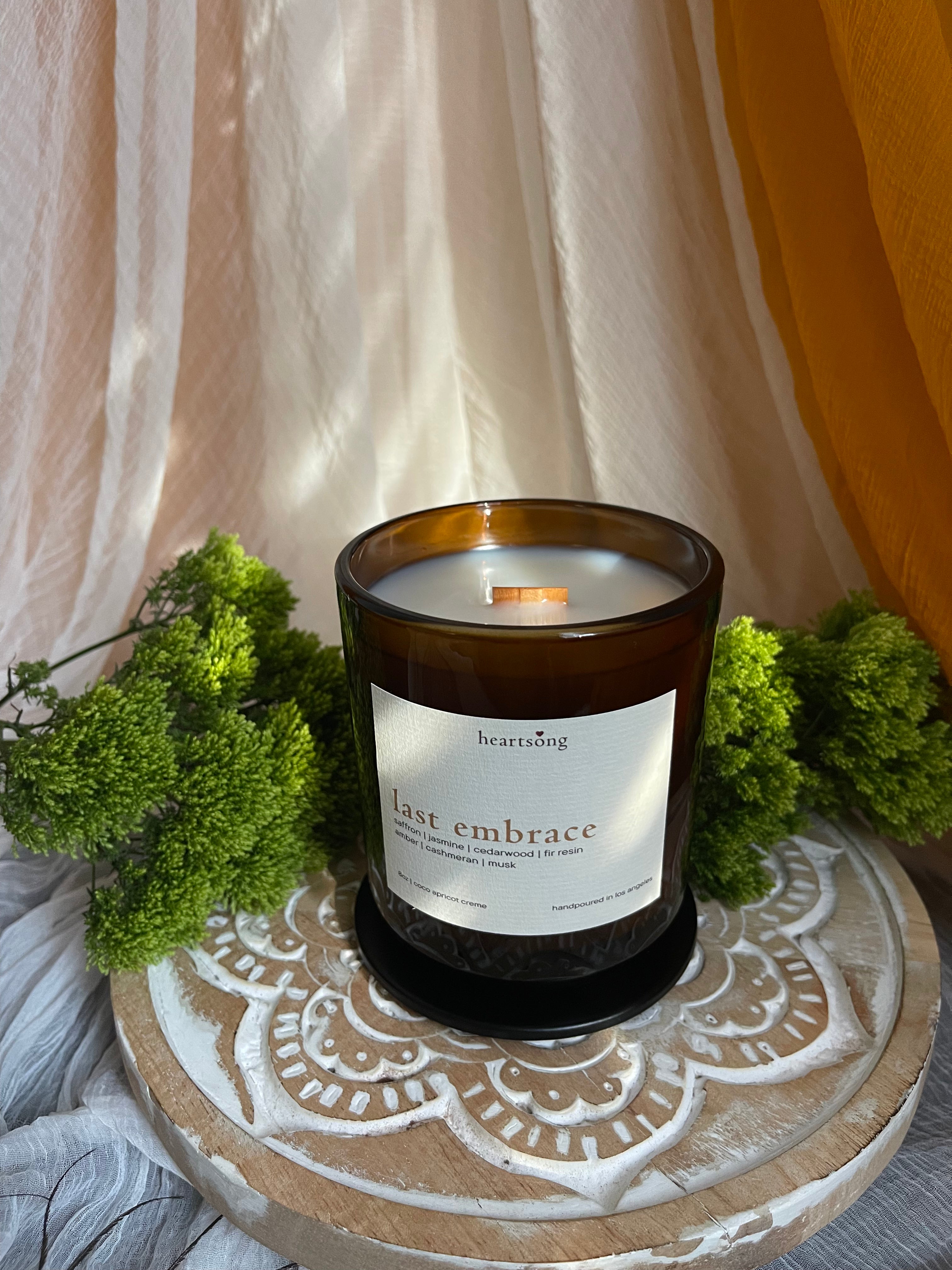 A heartstring brand soy candle in an amber glass jar with a white label reading 'last embrace', styled on a distressed wooden lotus stand with green foliage and draped yellow and cream fabric.