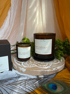 Two amber glass candles, one small and one larger with white labels reading ‘heartsong last embrace' are styled on a round wooden lotus board with greenery, draped fabric, and a peacock feather, alongside a dark brown product box.