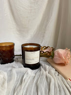 an amber glass jar candle with the name love in the morning on a white label sits on a white sheer fabric with an amber coffe mug, notebook, brass heart dish and peach coloured rose