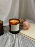a lit amber glass jar candle with the name love in the morning on a white label sits on a white sheer fabric with an amber coffe mug, notebook, and peach coloured rose