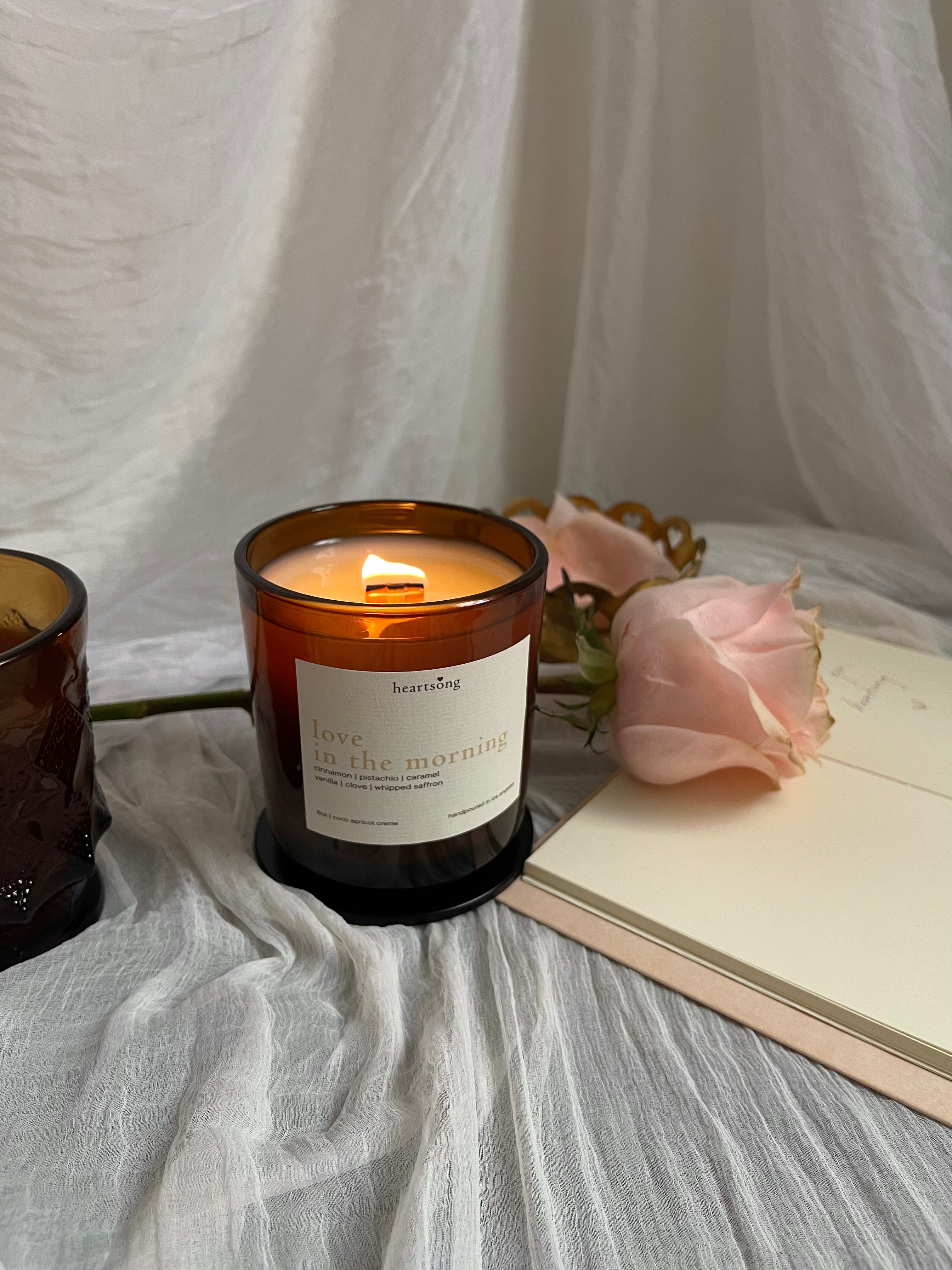 a lit amber glass jar candle with the name love in the morning on a white label sits on a white sheer fabric with an amber coffe mug, notebook, and peach coloured rose