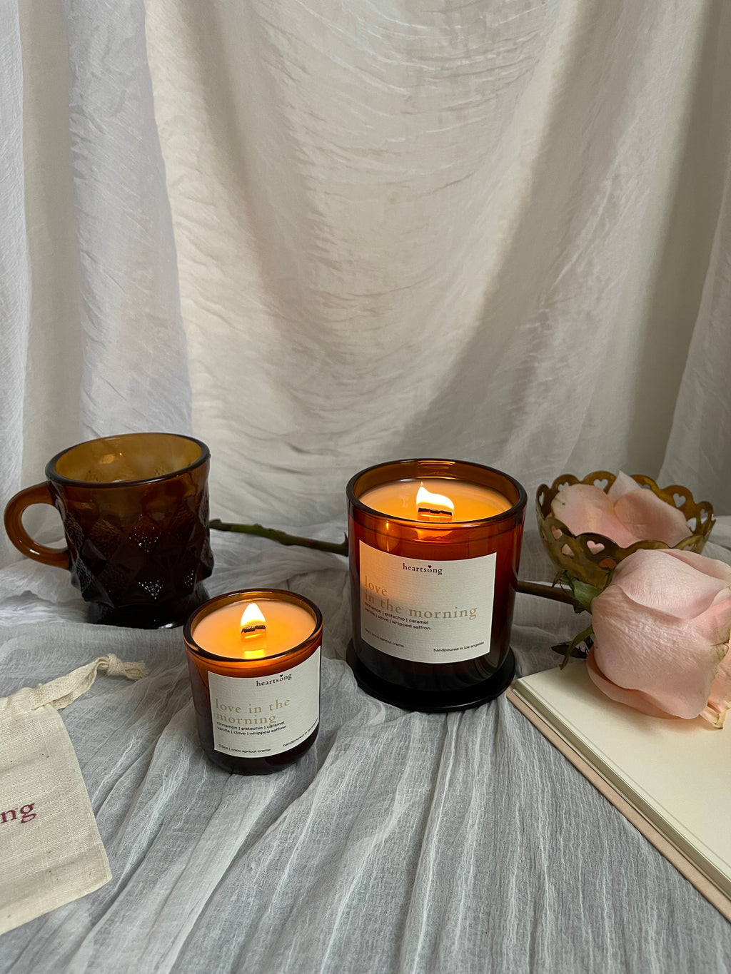 two lit amber glass jar candles with the name love in the morning on a white label sits on a white sheer fabric with an amber coffe mug, notebook, and peach coloured rose