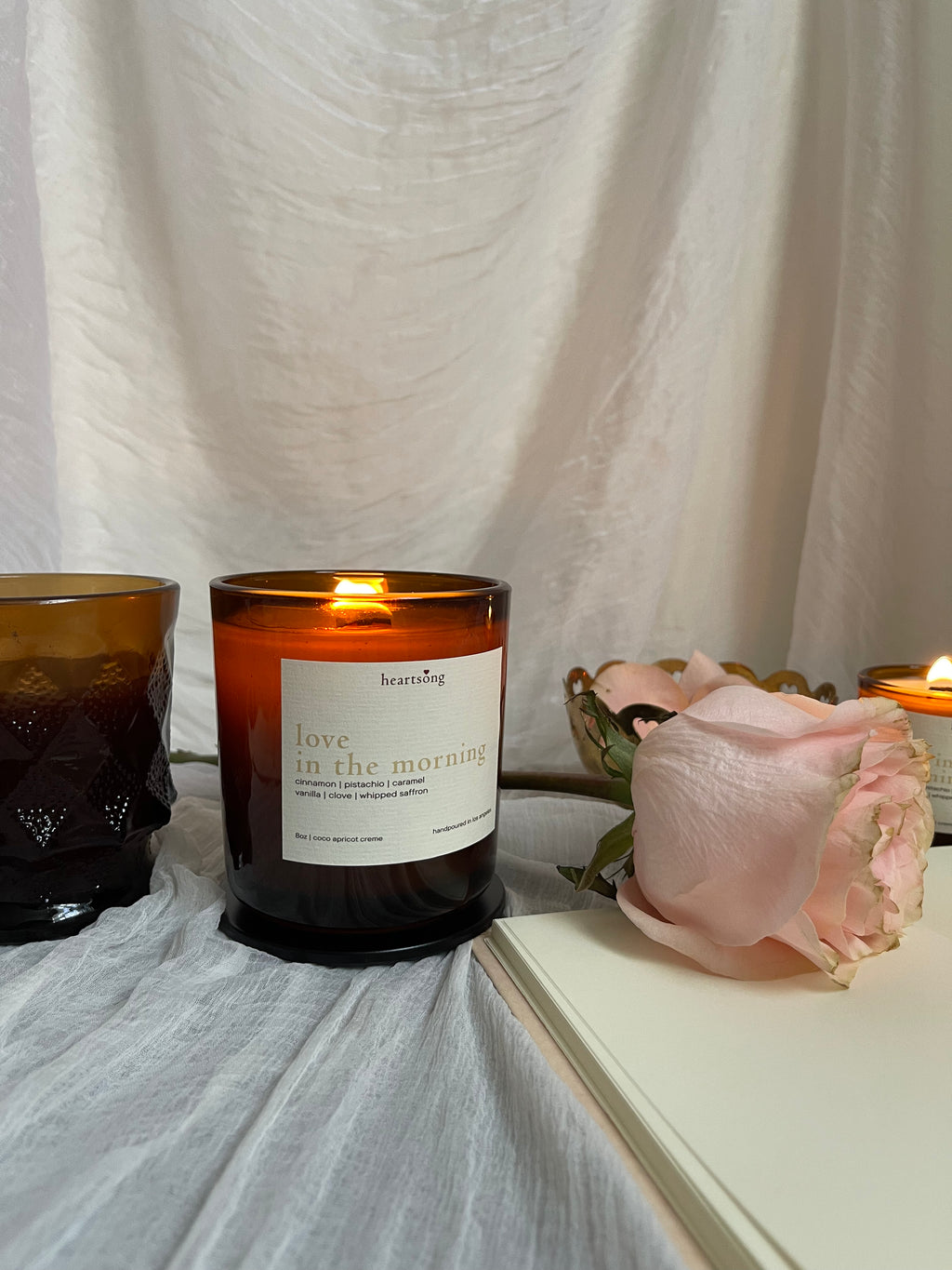a lit amber glass jar candle with the name love in the morning on a white label sits on a white sheer fabric with an amber coffe mug, notebook, and peach coloured rose