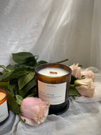 An amber glass jar candle labeled ‘heartsong' and ’swan song' sits on draped white fabric, surrounded by pink roses and green leaves. A partial second candle is visible at the left edge.