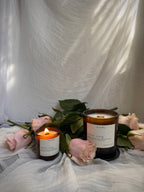 Two amber glass candles with white labels are styled with pink roses and green leaves against a draped white fabric backdrop. One candle is lit and the other sits on a dark brown saucer.