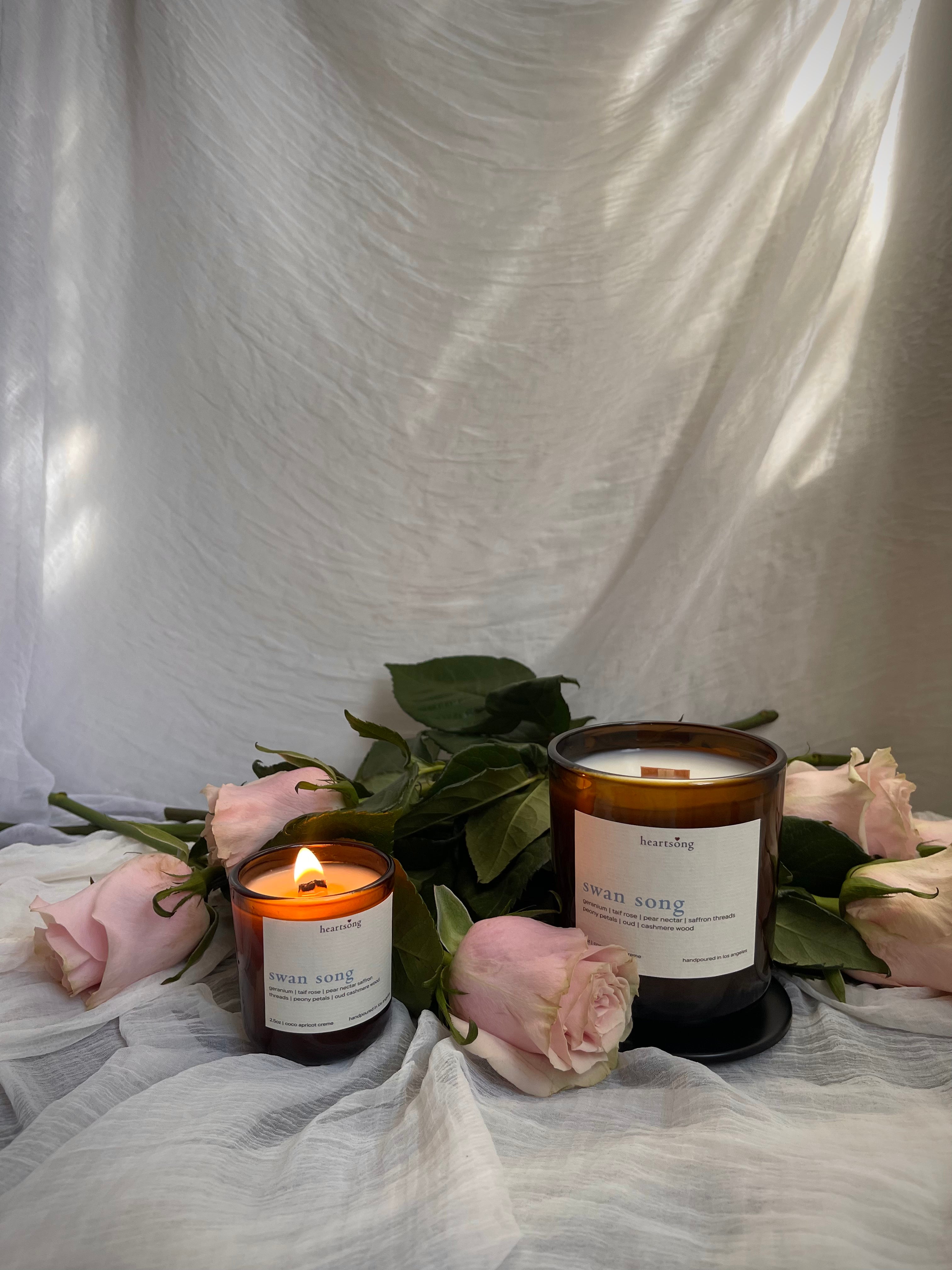 Two amber glass candles with white labels are styled with pink roses and green leaves against a draped white fabric backdrop. One candle is lit and the other sits on a dark brown saucer.