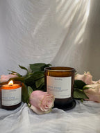 Two amber glass candles with white labels reading ‘heartsong' and 'swan song' are styled with pink roses and green leaves on draped white fabric. One candle is lit with a warm flame, and the background is a soft white fabric backdrop.