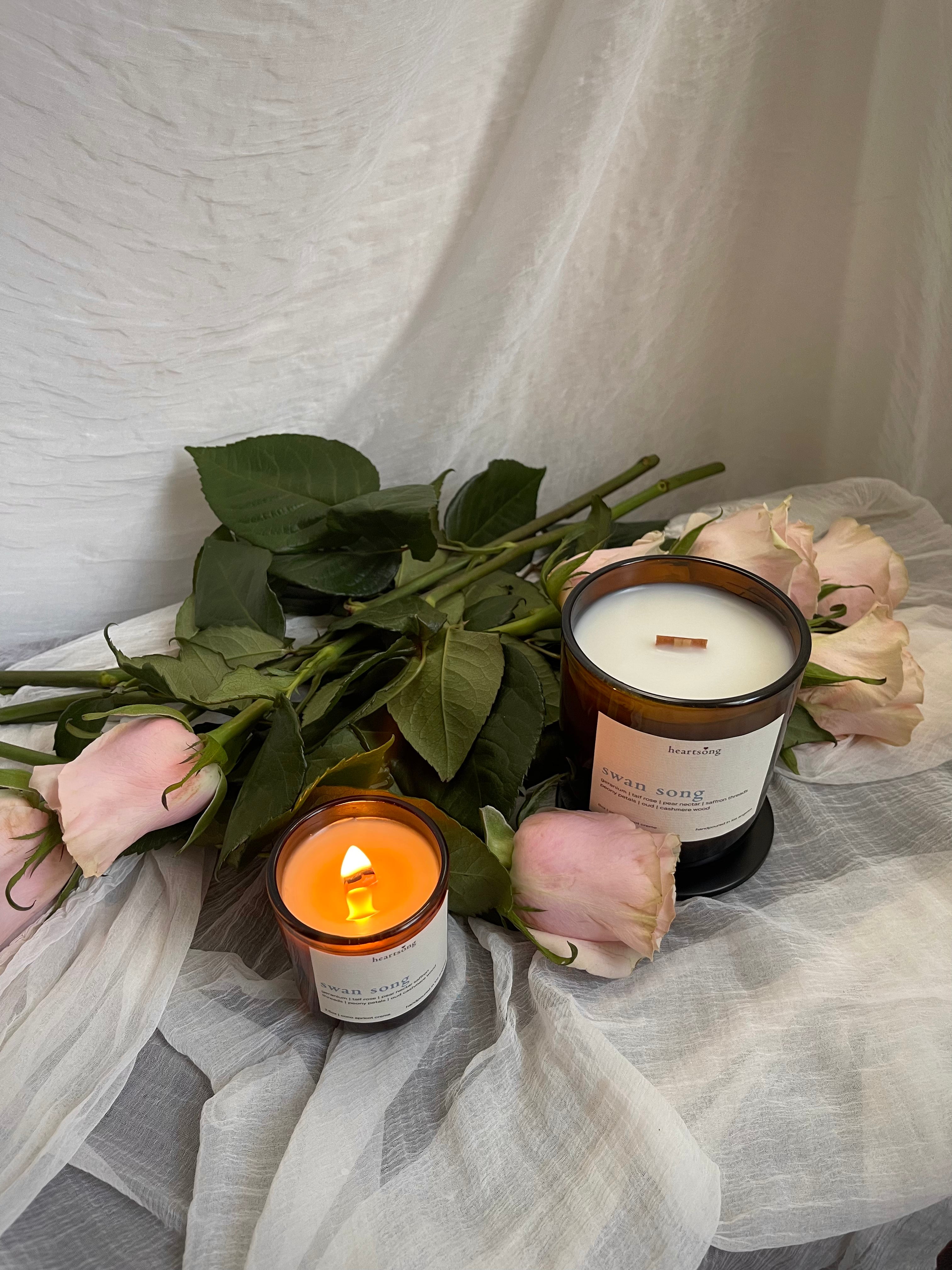 Two amber glass candles with white labels are styled with pink roses and green leaves on draped white fabric. One candle is lit and glowing, and the other is unlit.